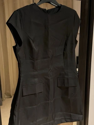 Svart kortärmad miniklänning - Stilren svart kortklänning med korta ärmar och rund hals. Klänningen har två dekorativa fickor framtill och en diskret dragkedja i ryggen. Perfekt för dig som gillar en enkel men chic look. Stor i storlek