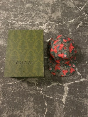 Gucci keps med jordgubbsmönster - Cool keps från Gucci med grå bas och ikoniskt GG-mönster, dekorerad med färgglada jordgubbar i rött och grönt. Kepsen har justerbar rem baktill och rosa innerfoder. Perfekt för dig som vill sticka ut med en unik accessoar.