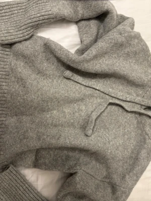 Grå stickad hoodie med huva - Mysig grå stickad hoodie med breda ribbade muddar vid ärmslut och nederkant. Hoodien har dragsko i huvan och är tillverkad i ett mjukt material som känns skönt mot huden. Perfekt för en avslappnad och chill stil.