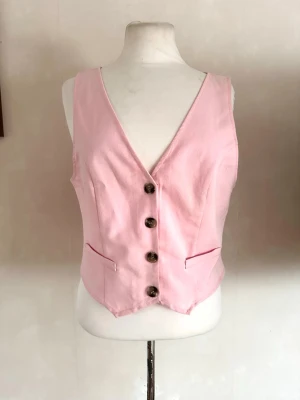 Ljusrosa väst med knappar - Snygg ljusrosa väst med djup V-ringning och fyra mörka knappar framtill. Västen har två dekorativa fickor och är figursydd för en schysst passform. Perfekt att styla över en t-shirt eller skjorta för en trendig look.