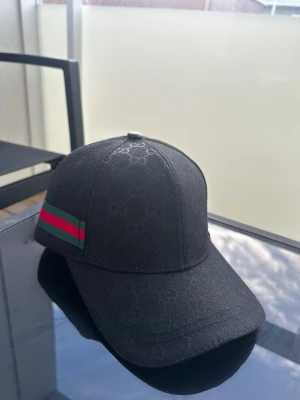 Svart keps från Gucci med GG-mönster - Säljer en svart keps från Gucci med diskret GG-monogram över hela kepsen och klassiska gröna och röda ränder på sidan. Kepsen är tillverkad i bomull och har justerbar passform. Perfekt för dig som vill ha en stilren och lyxig accessoar.