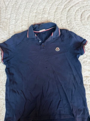 Marinblå Moncler pikétröja - Snygg marinblå pikétröja från Moncler med klassisk krage och två knappar framtill. Tröjan har korta ärmar och Moncler-logga på bröstet. Detaljer i vitt och rött på krage och ärmslut ger en sportig touch. Perfekt till jeans eller shorts. Den har en liten defekt på magen med inget som synns så mycket. Man får även med en gratis vara som synns på sista bilden🤯🤯🤯