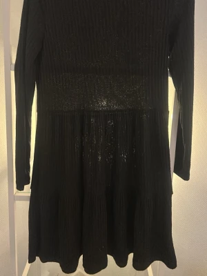 Svart glittrig klänning FB Sister - Snygg svart klänning från FB Sister med glittriga inslag. Klänningen har lång ärm, rund halsringning och är i kort modell. Materialet är stretchigt och skimrar i ljuset, perfekt för dig som vill sticka ut lite extra.