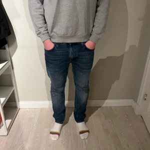 Blå slim fit jeans från Nudie Jeans - Säljer ett par blå slim fit jeans från Nudie Jeans med klassisk femficksdesign och snygga kontrastsömmar. Jeansen har normal midja och smal passform hela vägen ner. Perfekta till en avslappnad stil och tillverkade i mjukt denimtyg. De är i modellen lean dean i storlek 32x32