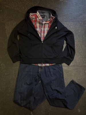 Marinblå hoodie från Polo Ralph Lauren - Snygg svart hoodie från Polo Ralph Lauren med dragkedja framtill och klassisk huva med dragsko. Ikoniska lilla loggan broderad på bröstet. Mjuk bomullsmix och två fickor framtill. Perfekt för en avslappnad och stilren look.