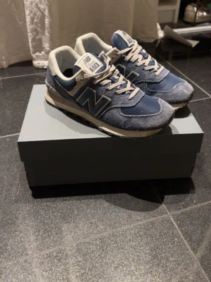 Blå New Balance 574 sneakers - Klassiska New Balance 574 sneakers i blått med grå och vita detaljer. Skorna har mocka- och meshmaterial, vita snören och den ikoniska N-loggan på sidan. Perfekta för en avslappnad och trendig stil. Nypris runt 1200