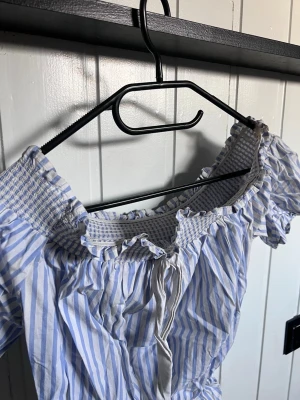 Blårandig offshoulder topp med volang - Söt offshoulder topp med blå och vita ränder, volangkant och smockad resår upptill. Knytband framtill och korta puffärmar ger en romantisk vibe. Perfekt för varma dagar och sommarhäng.