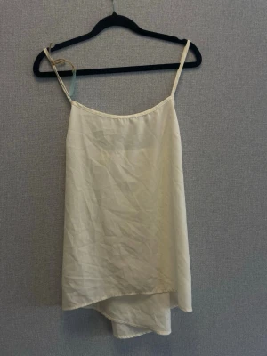 Beige linnetopp med asymmetrisk fåll - Säljer en beige linnetopp med tunna axelband och asymmetrisk fåll. Toppen har en enkel och stilren design som passar perfekt till jeans eller kjol. Materialet känns lätt och luftigt, perfekt för varma dagar.