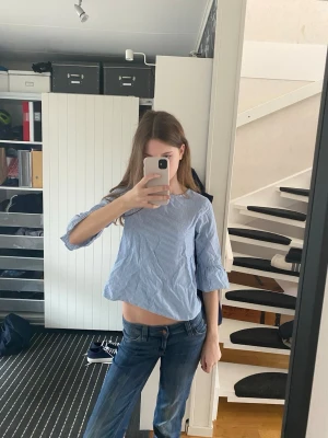 Blårandig blus med trumpetärm - Snygg blårandig blus med vita smala ränder och trumpetärmar i trekvarts längd. Blusen har rak passform och rund halsringning. Tillverkad i ett lätt bomullsliknande material som känns fräscht och luftigt. Perfekt till jeans för en avslappnad men trendig look.