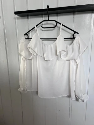 Vit offshoulder blus med volang - Trendig vit offshoulder blus med bred volang runt axlarna och tunna axelband. Blusen har långa ärmar med resår och volangdetalj vid ärmslut. Tillverkad i ett lätt och luftigt tyg som ger en romantisk känsla.