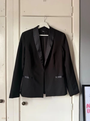 Svart kavaj med satindetaljer från bikbok - Stilren svart kavaj från bikbok med blanka satindetaljer på slag och fickor. Klassisk rak passform och helsvart design som ger en elegant look. Perfekt till att lyfta vilken outfit som helst.