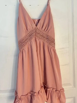 Rosa kort klänning med spetsdetaljer - Söt rosa kortklänning med tunna axelband och v-ringning. Klänningen har fina spetsdetaljer under bysten och volangkant nedtill. Perfekt för sommaren eller utomlands. Varan är ej använd endast testad.