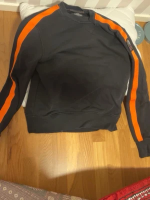 Parajumpers sweatshirt - Cool svart sweatshirt från Napapijri med orange ränder längs ärmarna och rund hals. Tröjan har en avslappnad passform och ribbade muddar vid ärmslut och nederkant. Perfekt för en sportig och trendig look. NYPRIS 1600kr