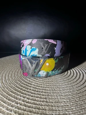 Färgglatt LV-bälte i skinn - Louis Vuitton skärp i svart skinn med färgglatt blommigtw mönster och stor silverfärgad LV-logga som spänne. Skärpet har inslag av lila, blått, gult och rosa i designen och är tillverkat i Spanien. Perfekt statement-accessoar för att lyfta din outfit.