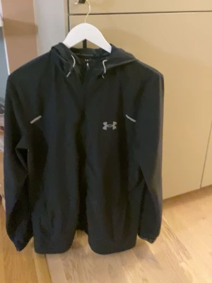 Svart vindjacka från Under Armour - Snygg svart vindjacka från Under Armour med huva och dragsko. Jackan har reflekterande detaljer och logga på bröstet samt ett stort Under Armour-tryck på ryggen. Tillverkad i lätt och tåligt material, perfekt för blåsiga dagar. Priset är ej hugget i sten
