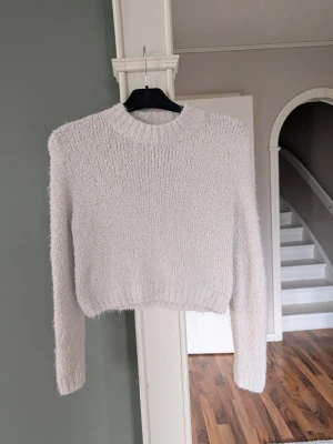 Ljusgrå fluffig stickad tröja - Mysig ljusgrå stickad tröja med fluffig textur och rund hals. Tröjan har långa ärmar och ribbstickade muddar vid hals, ärmslut och nederkant. Perfekt för kyliga dagar och enkel att matcha med olika outfits.