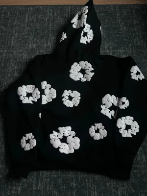 Svart hoodie från Denim Tears - Säljer en svart hoodie från Denim Tears med stora vita blommönster över hela plagget. Hoodien har en klassisk huva och är gjord i mjukt bomullsmaterial. Perfekt för dig som vill sticka ut med en unik och trendig design.