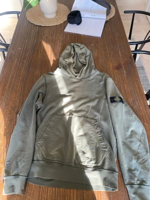 Olivgrön hoodie från Stone Island - Säljer en olivgrön hoodie från Stone Island med klassisk patch på vänster ärm. Hoodien har en stor känguruficka framtill och ribbade muddar. Tillverkad i mjuk bomull och har en avslappnad passform med huva.