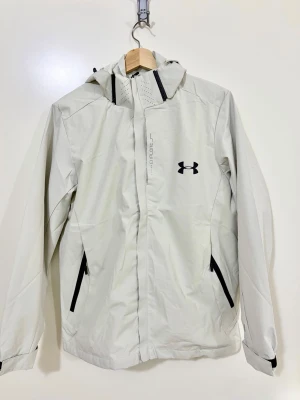 Vit vindjacka från Under Armour S/M storlek  - Snygg vit vindjacka från Under Armour med svart logga på bröstet och svarta dragkedjor på fickorna. Jackan har huva, hel dragkedja framtill och meshfoder för extra ventilation. Perfekt för blåsiga dagar och sportiga outfits.