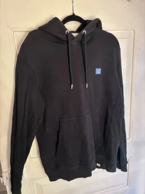 Svart hoodie från Les Deux - Snygg svart hoodie från Les Deux med klassisk känguruficka och justerbar huva med svarta snören. Liten blå logga på bröstet ger en clean look. Tillverkad i mjukt bomullsmaterial som är skönt att ha på sig hela dagen. Passar snarare S.