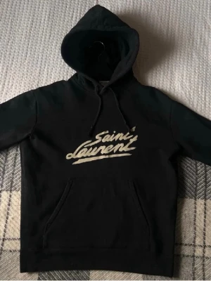 Svart Saint Laurent hoodie - Snygg svart hoodie från Saint Laurent med stor vit logga broderad på bröstet. Klassisk huva med dragsko och känguruficka framtill. Perfekt för dig som gillar streetwear och vill ha något stilrent men ändå trendigt.