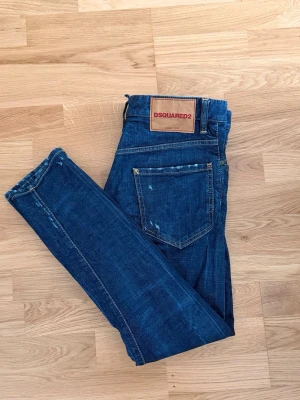 Dsquared2 Jeans - Size 46 IT (S/M) - Selling a pair of authentic and very stylish DSQUARED2 jeans. These jeans are a dark blue wash with factory distress, minor tears, and authentic wear details on the pocket edges and hems for that perfect, lived-in designer look. Feature a classic button fly and the signature red Dsquared2 tab. Made in Italy. High-quality denim with a premium feel. Brand: Dsquared2 Size: Italian 46 (Corresponds to approx. a size S or small M. Please check the fit of this model). Condition: Only tried 