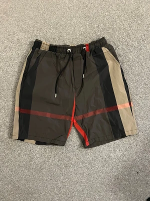 Från burrbery badshorts med snörning - Snygga badshorts med klassiskt rutigt mönster i svart, beige och rött. Shortsen har snörning i midjan och sidofickor. Perfekta för sommaren och strandhäng.