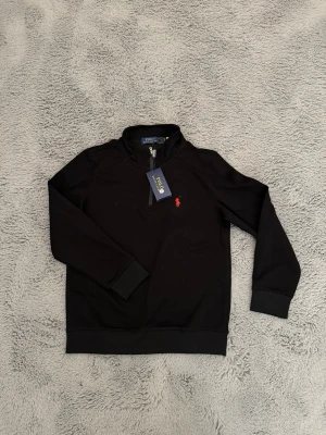 Svart half-zip hoodie Polo Ralph Lauren - Snygg svart half-zip hoodie från Polo Ralph Lauren med klassisk röd logga broderad på bröstet. Tillverkad i mjukt material med ribbade muddar och hög krage. Perfekt för dig som vill ha en stilren och bekväm hoodie med premiumkänsla. (Ingår påse)