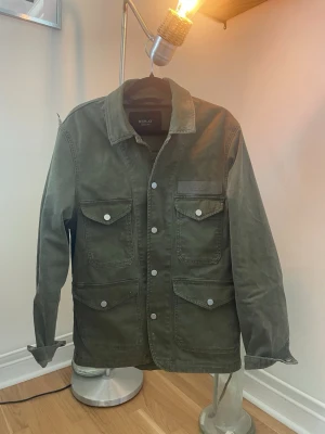 Olivgrön overshirt från Replay - Stilren olivgrön overshirt från Replay med fyra stora fickor framtill och tryckknappar. Overshirten har klassisk krage och ett tygmärke med Replay-loggan på bröstet. Tillverkad i ett slitstarkt bomullstyg som ger en avslappnad look.