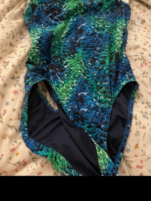 Mönstrad nike baddräkt  - Säljer en snygg nike baddräkt med abstrakt mönster i blått, grönt och svart. Klassisk modell med hög skärning vid benen och rundad halsringning. Materialet känns stretchigt och bekvämt, perfekt för simning eller strandhäng.