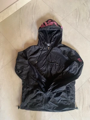 Svart vindjacka med rosa detaljer - Snygg svart vindjacka från Risky med dragkedja, huva och justerbar nederkant. Jackan har en rosa logga på ärmen och rosa mönster på insidan av huvan. Tillverkad i lätt och tåligt syntetmaterial, perfekt för blåsiga dagar.