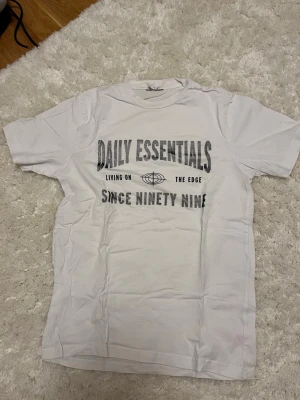 Vit Daily Essentials t-shirt - Vit t-shirt från Daily Essentials med svart tryck framtill där det står 'Living on the edge since ninety nine' och en grafisk detalj. Klassisk passform och rund hals. Perfekt för en chill och avslappnad stil.