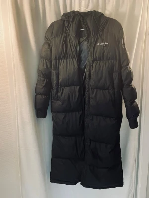 Svart lång dunjacka från Columbia - Säljer en svart lång dunjacka från Columbia med huva och dragkedja hela vägen ner. Jackan har en puffig design med breda kanaler och logga på bröstet. Perfekt för kalla vinterdagar och riktigt varm.