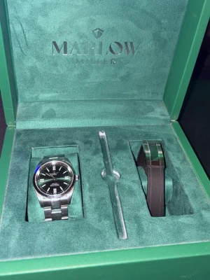 Silverfärgad klocka från Marlow Miller - Stilren klocka från Marlow Miller med silverfärgad länk och svart urtavla. Kommer i original grön box och extra armband. Klassisk rund form och tydliga markeringar på urtavlan. Perfekt accessoar för dig som gillar en clean look.