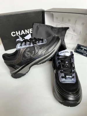 Svarta sneakers från Chanel - Snygga svarta sneakers från Chanel med metallicdetaljer och klassisk logga på sidan. Skorna har en chunky sula, svarta skosnören och materialmix av mesh, läder och syntet. Perfekta för dig som vill ha en exklusiv och trendig look.