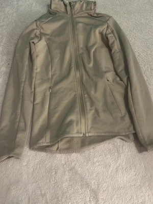 Beige vindjacka med huva - Snygg beige vindjacka med huva och hel dragkedja framtill. Jackan har två sidofickor med dragkedja och är tillverkad i ett lätt syntetmaterial. Perfekt för lager-på-lager och har en clean, sportig look med lång ärm och enkel design. 