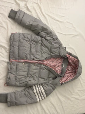 Grå thom browne puffer jacka - Snygg grå pufferjacka med huva och vita ränder på ena ärmen. Jackan har ribbade muddar, dragkedja och tryckknappar framtill samt ett rödvitt randigt innerfoder. Perfekt för kalla vinterdagar och riktigt trendig look. Thom browne jacka men tyvärr saknas en av knapparna men det är lätt fix att föra till någon som fixar det eller fixa det själv(märks dock knappt att den saknas)