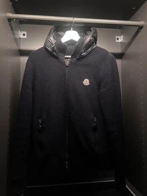 Mörkblå stickad jacka från Moncler - Snygg mörkblå stickad jacka från Moncler med dragkedja framtill och två fickor med dragkedja. Jackan har en quiltad huva i blankt material och Moncler-logga på bröstet. Perfekt för kyliga dagar och ger en exklusiv känsla till din outfit. Skick är 9/10 sjukt svårt o få tag i denna modellen! 100% äkta kan fixa kvitto