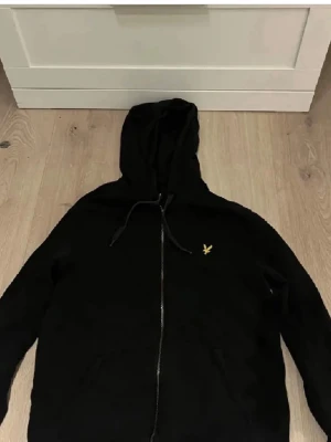 Svart hoodie med dragkedja Lyle & Scott - Säljer en svart hoodie från Lyle & Scott med dragkedja framtill och klassisk gul logga på bröstet. Hoodien har huva med dragsko och två fickor framtill. Perfekt för dig som gillar enkel och stilren streetwear.