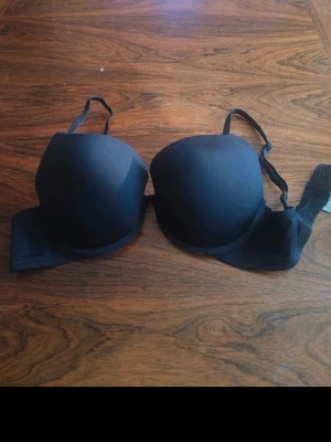 Svart push-up bh med justerbara band - En enkel och stilren svart push-up bh med vadderade kupor och justerbara axelband. Bh:n har slät yta utan synliga detaljer och stängs med hyskor bak. Perfekt för dig som vill ha extra lyft och stöd.
