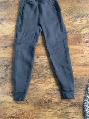 Nike tech - Snygga mörkgråa mjukisbyxor från Nike med smal passform och ribbade muddar vid bensluten. Byxorna har dragkedjeförsedd ficka på ena sidan och klassisk Nike-logga tryckt på benet. Tillverkade i mjukt och bekvämt material, perfekta för chill eller träning.