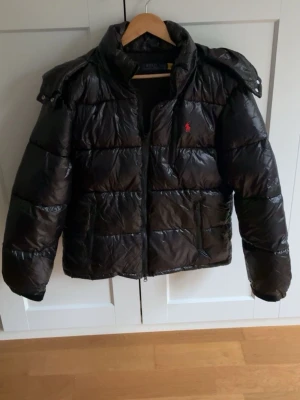 Svart pufferjacka från Polo Ralph Lauren - Snygg svart pufferjacka från Polo Ralph Lauren med blank finish och röd logga på bröstet. Jackan har avtagbar huva, dragkedja framtill och två sidofickor med dragkedja. Perfekt för kalla vinterdagar och riktigt trendig look.