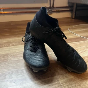 Svarta Nike fotbollsskor med socka - Säljer ett par svarta Nike fotbollsskor med högre strumpkrage för extra stöd runt ankeln. Skorna har snörning, diskret Nike-logga i vitt på sidan och är tillverkade i syntetmaterial med struktur för bättre bollkontroll. Perfekta för dig som vill ha stilrena och funktionella skor på planen.