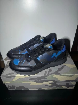 Blå camo sneakers från Valentino - Snygga sneakers från Valentino med blå och svart camo-mönster. Skorna har svarta snören, svart sula med rutmönster undertill och detaljer i både mocka och läder. Perfekt för dig som vill sticka ut med en unik och trendig look.