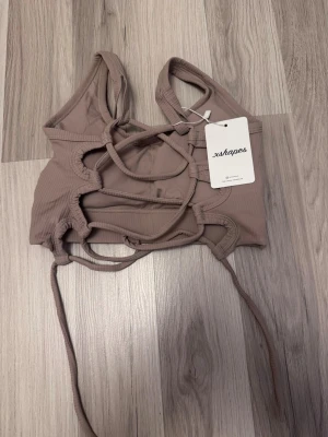 Beige ribbad linnetopp från xshapes - Trendig beige linnetopp från xshapes med ribbad struktur och snygga snörningsdetaljer framtill. Toppen har breda axelband, croppad passform och ett litet vitt xshapes-tryck på bröstet. Perfekt för att lyfta din outfit med en edgy touch.