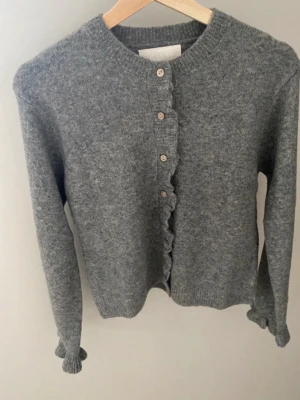 Grå kofta med volangkant - Mysig grå kofta med volangkant längs knappraden och vid ärmsluten. Rund halsringning och ribbstickade muddar. Stängs med små knappar framtill. Perfekt för lager på lager och enkel att styla.