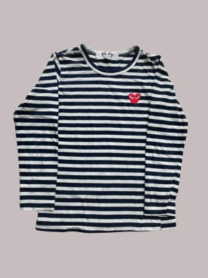 Randig långärmad topp Comme des Garçons - Säljer en klassisk långärmad topp från Comme des Garçons Play med marinblå och vita ränder samt det ikoniska röda hjärtat på bröstet. Tillverkad i mjuk bomull och har en avslappnad passform. Säljer för jättebra pris (original pris 1000-1500kr) defekt finns på sista bilden