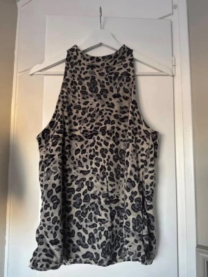 Leopardmönstrad ärmlös topp - Snygg ärmlös topp med leopardmönster i grå och svarta toner. Toppen har hög halsringning och loose passform, perfekt för att sticka ut lite extra. Materialet känns mjukt och följsamt mot huden.