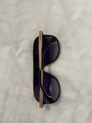 Svarta Carrera solglasögon med guld - Snygga solglasögon från Carrera med svart båge och guldfärgade skalmar. Glasen är mörka och har en klassisk aviatorform som ger en cool vibe. Perfekta för att lyfta din stil under soliga dagar.