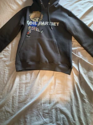 Svart hoodie från Goil partiet - Svart hoodie med huva och dragsko från Goil partiet. Framsidan har ett stort tryck med en färgglad figur och texten 'GOIL PARTIET' i glittriga stenar. Klassisk känguruficka framtill och ribbade muddar. Perfekt för dig som gillar statement-plagg och streetwear. (LIMITED EDITION)Priset kan diskuteras.